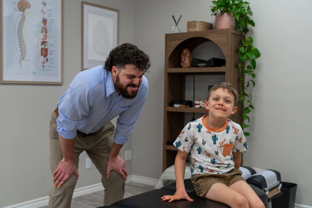 Smiling child sitting on chiropractic table with chiropractor observing, symbolizing a positive and supportive environment for children with behavioral challenges.