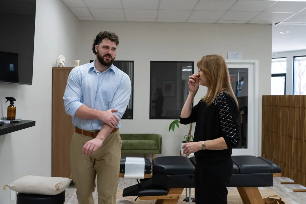 Dr. Korrin Taylor speaks with a female patient beside an adjustment table in a clinic room.