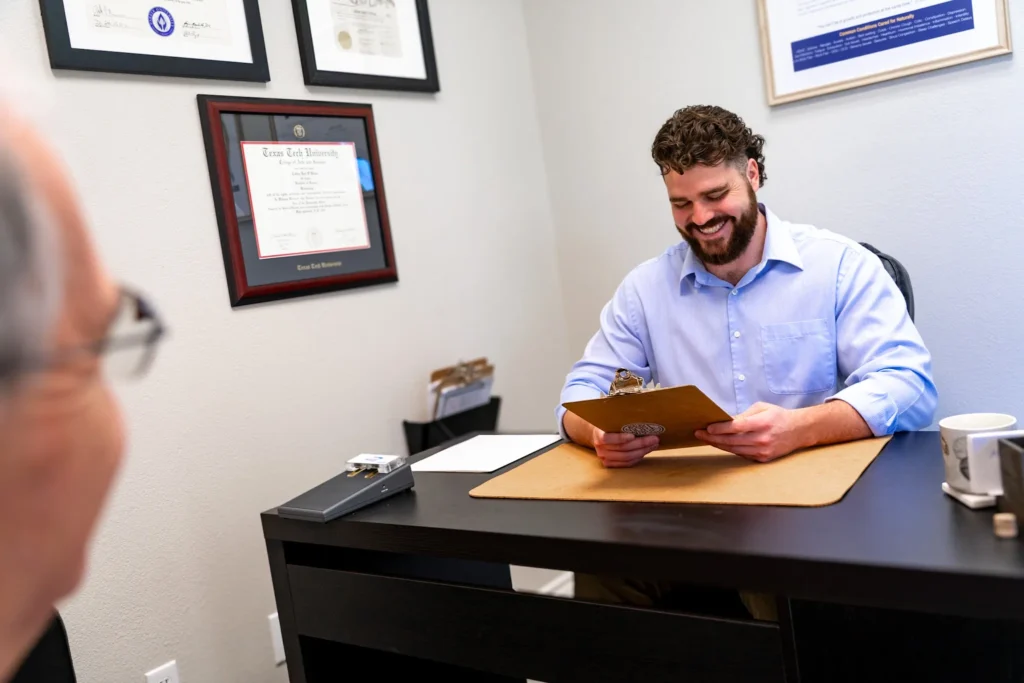 Dr. Korrin Taylor sits at his desk smiling while holding a clipboard during a patient consultation.