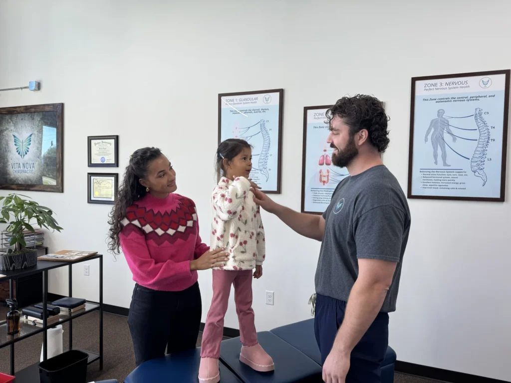 Dr. Korrin Taylor examines a young girl standing on a chiropractic table while her mother supports her.