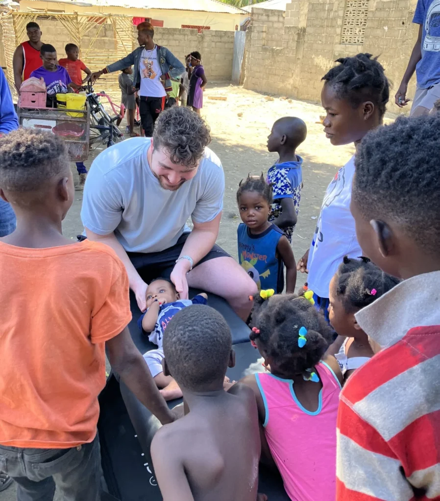 Dr. Korrin Taylor gently supports an infant on a portable table outdoors surrounded by smiling children.