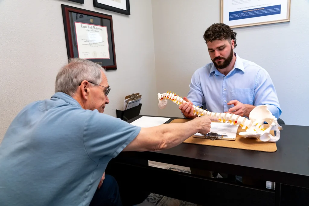 Dr. Korrin Taylor reviews an anatomical spine model with a patient during a consultation at his desk.
