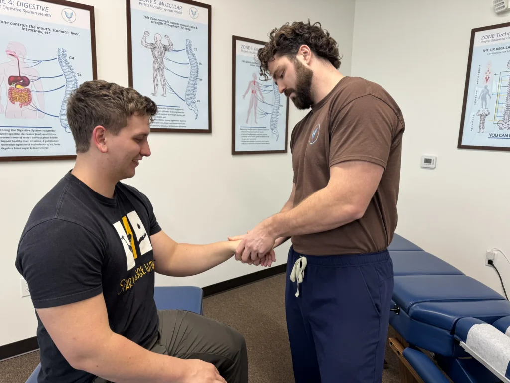 Dr. Korrin Taylor examines a seated patient's wrist and hand in a treatment room.