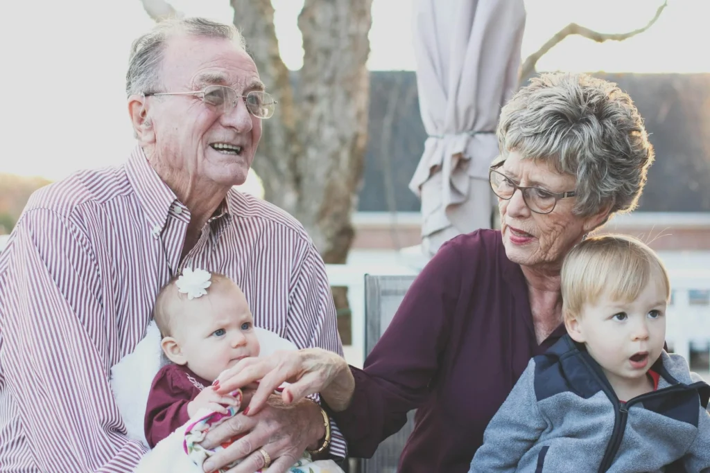 An older couple smiling while sitting outdoors together holding a baby and a young toddler.