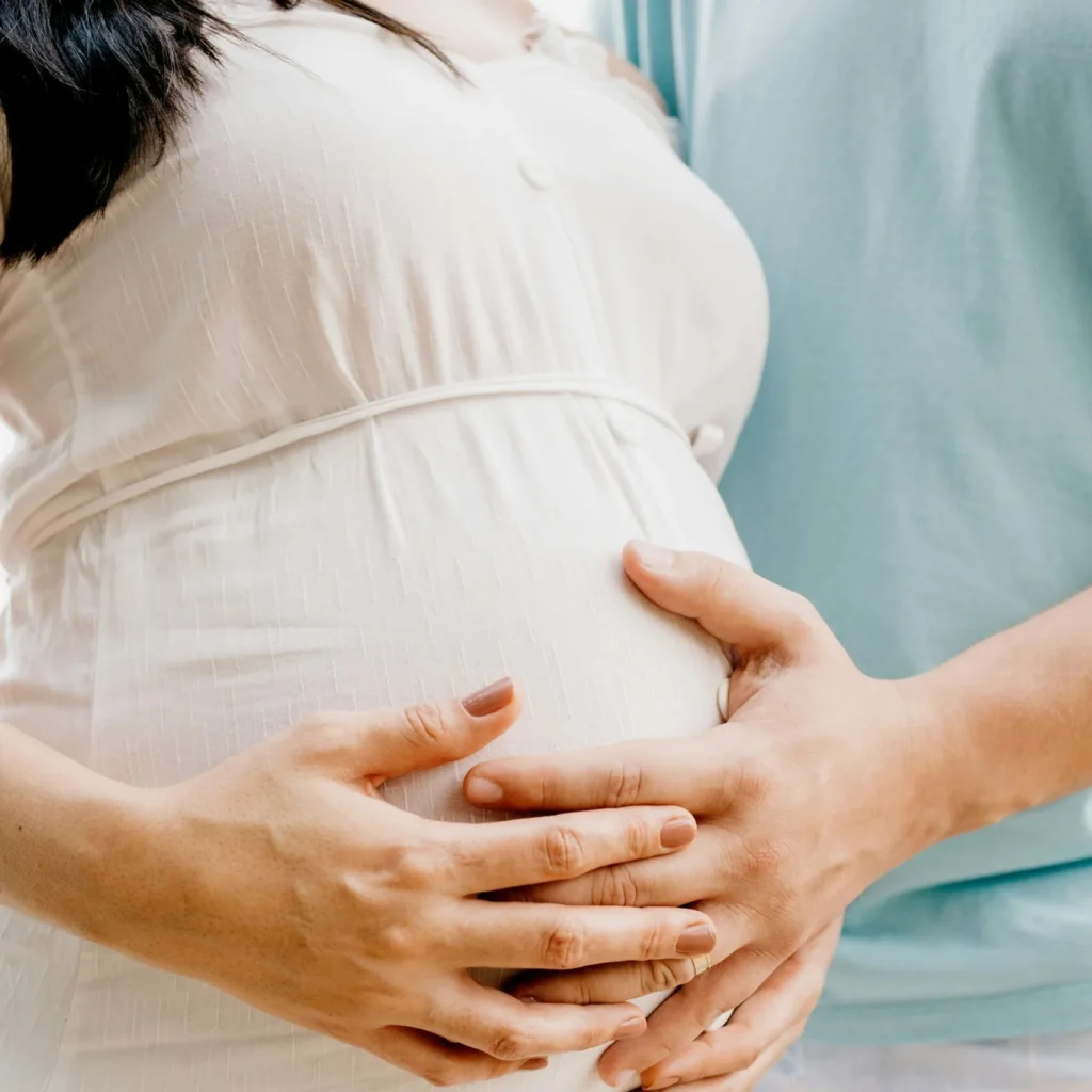 Close-up of a pregnant woman in a white dress with her and her partner's hands resting gently on her belly.