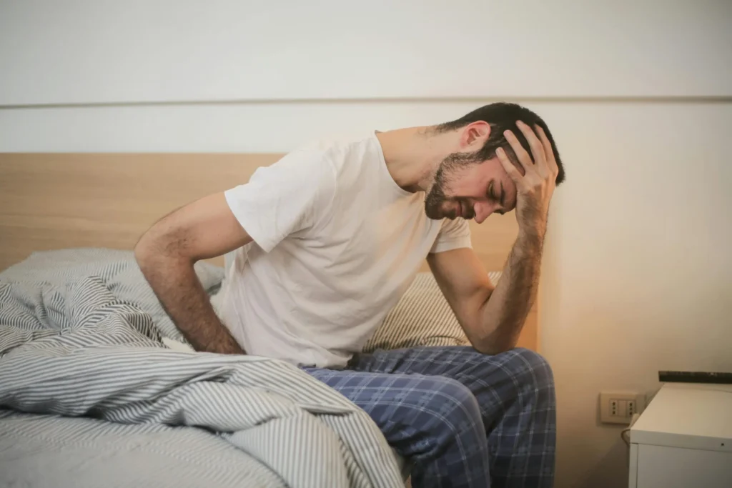 A man in pajamas sits on the edge of a bed holding his lower back and forehead in visible discomfort.