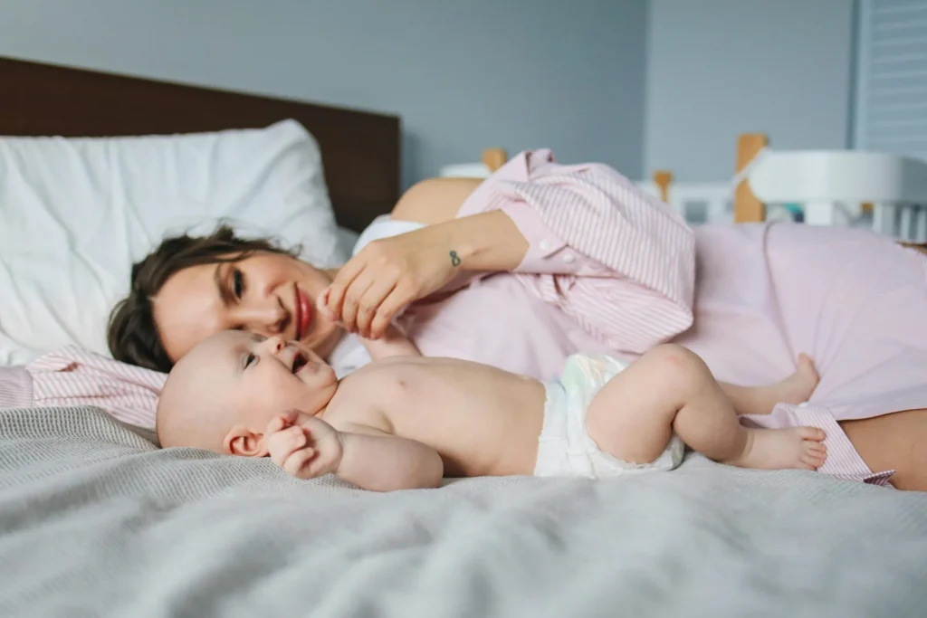 Mother smiling while lying on a bed next to her playful infant in a diaper.
