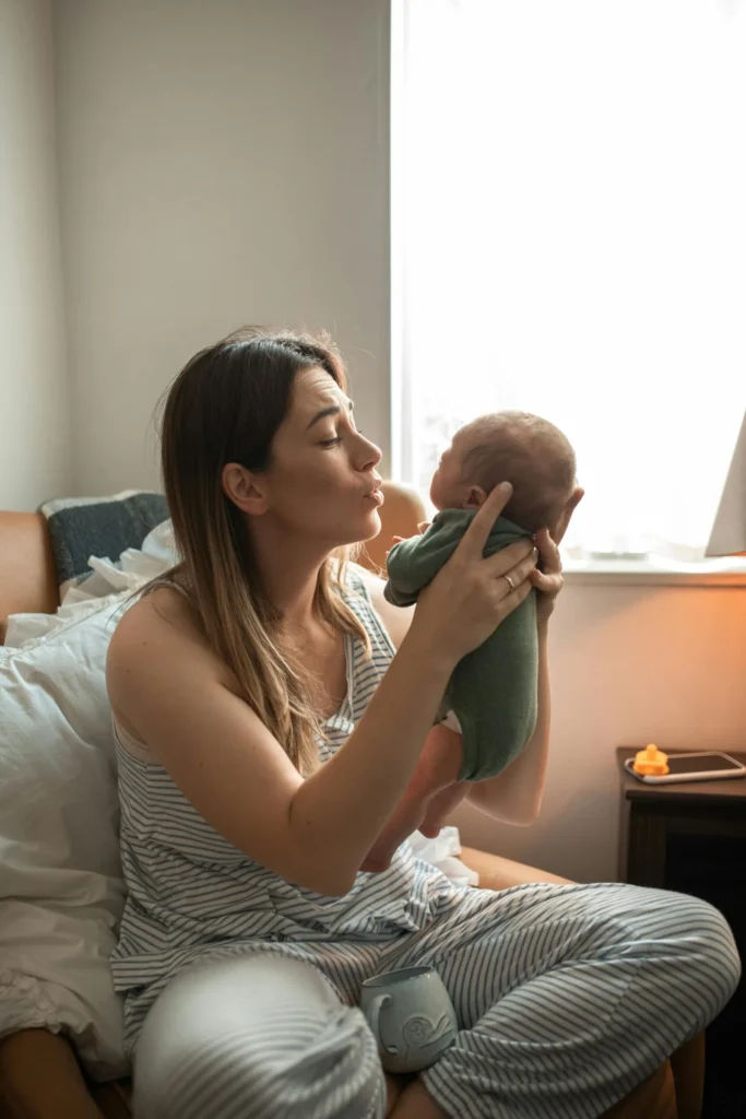 Mother sitting in an armchair holding up her newborn baby near a bright window.