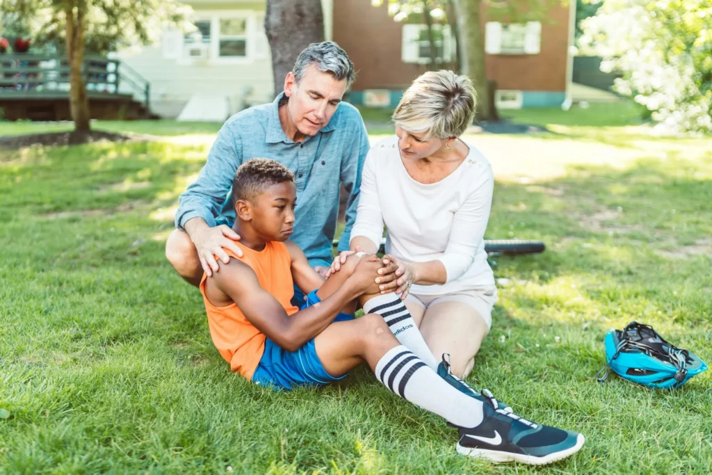A man and woman comfort a boy holding his knee while sitting on a grassy lawn with a bicycle nearby.