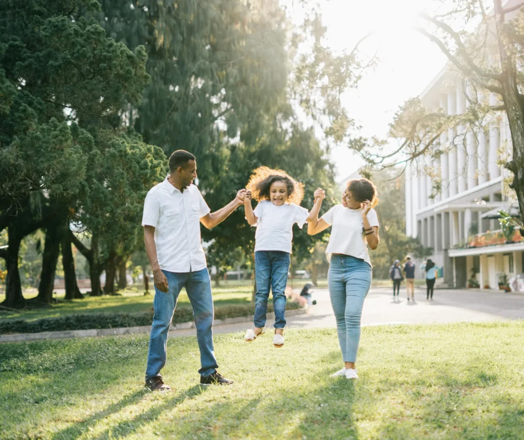 Parents holding hands with their young daughter and lifting her into the air while walking across a sunny lawn.