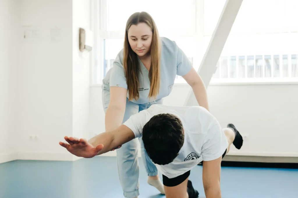 A practitioner in scrubs guides a man performing a core stabilization exercise on a blue floor in a bright clinic.
