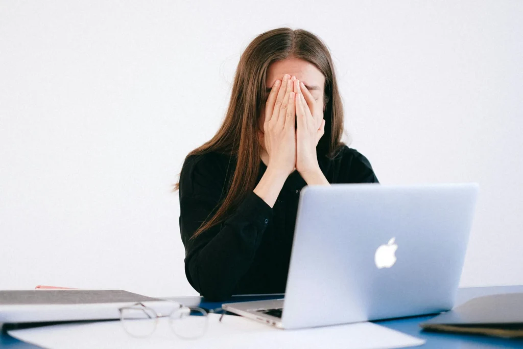 Woman sitting at a desk covering her face with both hands in front of an open laptop.