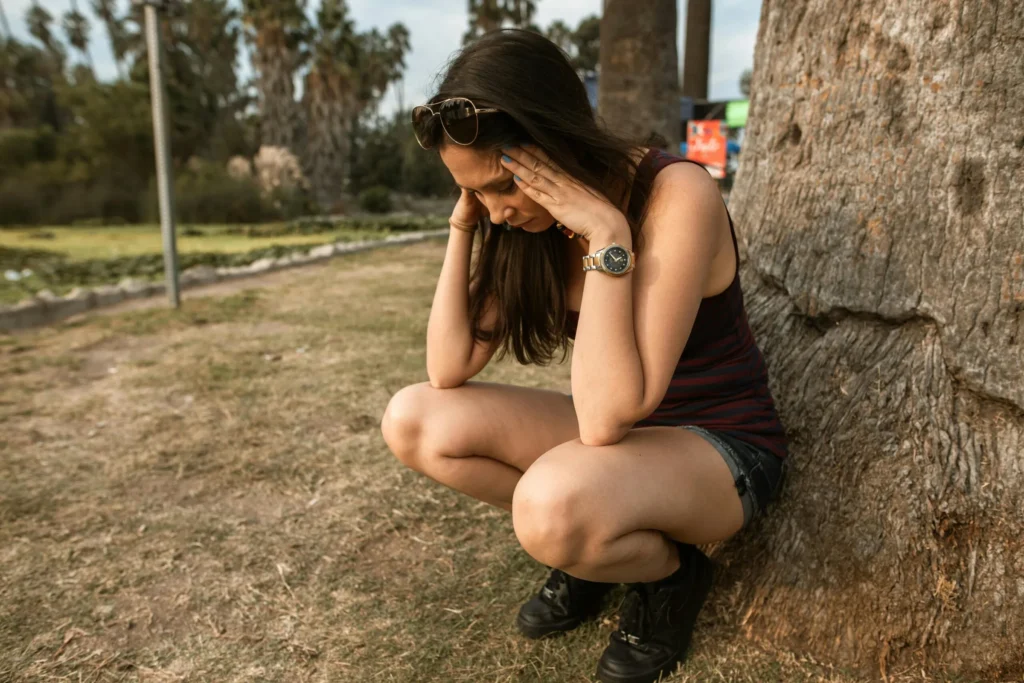 Woman crouching next to a tree trunk outdoors with both hands resting against her temples.