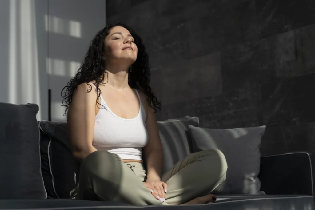 A woman sitting cross-legged on a couch with her eyes closed, resting in the sunlight.